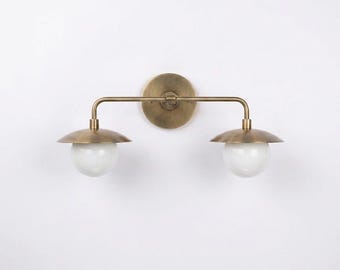 May include: A two-light wall sconce featuring a circular brass backplate, a horizontal brass arm, and two downward-facing brass shades. Each shade is paired with a spherical white glass globe bulb, creating a balanced and symmetrical lighting fixture.
