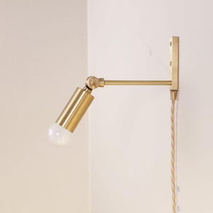 May include: A brass wall-mounted lamp with an adjustable arm and a cylindrical light fixture. The lamp has a clear bulb and a braided cord. The fixture is mounted on a white wall.
