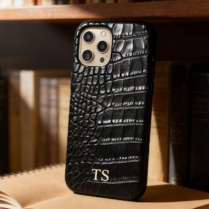 May include: Black phone case with a crocodile skin texture, featuring a gold camera lens surround and the initials "TS" in gold. The case is displayed on an open book, with a backdrop of bookshelves.