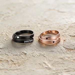 Puede incluir: Dos anillos de estilo envolvente, uno negro mate y otro oro rosa. El anillo negro está grabado con "I [símbolo de corazón]" y "you [símbolo de infinito]". El anillo de oro rosa está grabado con "John [símbolo de corazón]" y "[símbolo de corazón] Luna".