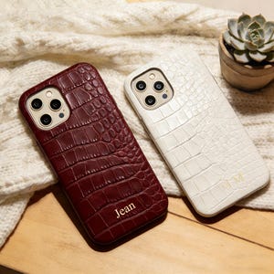 Puede incluir: Dos fundas para teléfono con textura de piel de cocodrilo. Una es de color burdeos oscuro con el nombre "Jean" impreso. La otra es blanca con las iniciales "MM" impresas. Ambas fundas tienen un recorte para la cámara.