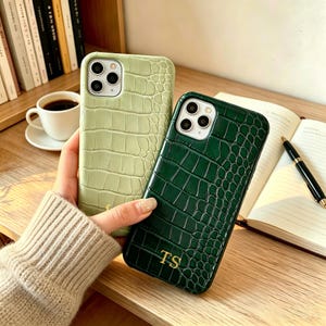 Puede incluir: Dos fundas para smartphone con un patrón de textura de piel de cocodrilo. Una funda es de color verde salvia claro y la otra es de color verde bosque profundo con las iniciales TS grabadas en oro en la parte inferior. Ambas tienen triple cámara.