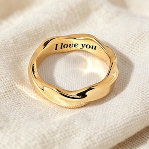 Peut inclure: Une bague dorée avec une surface irrégulière et ondulée. L'intérieur de l'anneau présente le texte gravé en noir "I love you" dans une police serif. La bague possède une finition polie et réfléchissante qui capte la lumière sur ses courbes.
