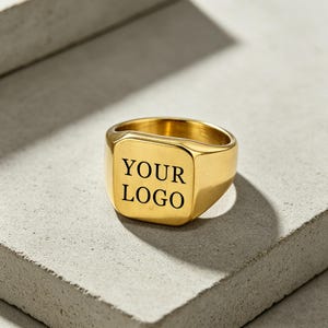 May include: A gold-toned signet ring featuring a flat, square-shaped face with the text YOUR LOGO engraved in black lettering. The ring has a polished metallic finish and a smooth, rounded band that tapers towards the back of the piece.