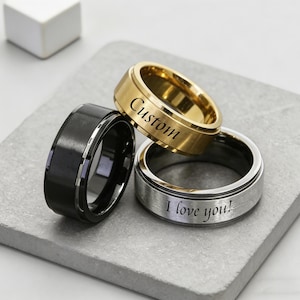 May include: Three metal rings with a smooth finish. One ring is solid black, one is gold-toned with the word Custom engraved on the surface, and one is silver-toned with the engraved text I love you! visible on the outer band of the ring.