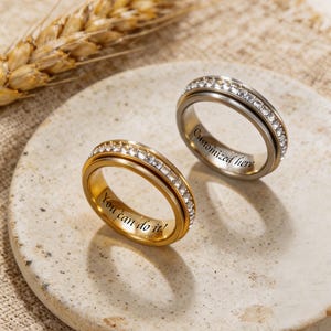 Pode incluir: Dois anéis, um dourado e outro prateado, cada um com uma faixa de pedras transparentes. O anel dourado tem a inscrição "You can do it!" e o anel prateado "Customized here". Os anéis são exibidos em uma superfície texturizada esbranquiçada.