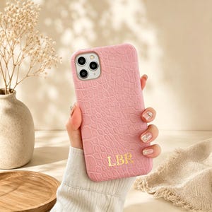 May include: A light pink phone case with a textured crocodile skin pattern and the gold embossed letters LBR at the bottom. The case features a cutout for a triple-lens camera system and is designed to fit a smartphone securely and protectively.