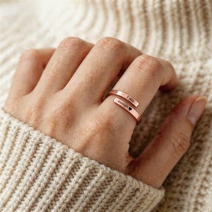 以下が含まれることがあります： ローズゴールド調のオープンデザインのラップリング。上端には「I am」、下端には「enough」という文字が刻印されており、その後に小さな黒いハートのシンボルが金属表面に施されています。