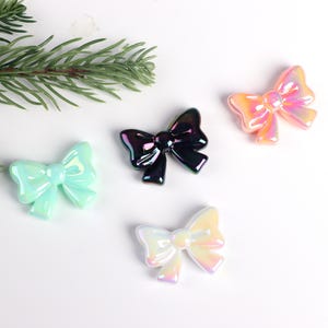 May include: Four iridescent plastic bows in different colors: light blue, black, light pink, and white. The bows are arranged on a white surface.