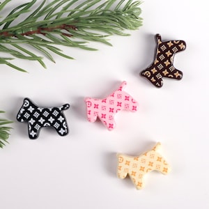 May include: Four dog-shaped decorative embellishments in black, pink, brown, and cream. Each dog features a repeating pattern of a four-petal flower design. A sprig of green pine needles is in the upper left corner.