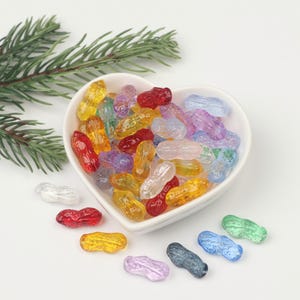 May include: A heart-shaped bowl filled with colourful glass peanut-shaped beads. The beads are in various shades of red, orange, yellow, green, blue, purple, and clear. Some beads are scattered around the bowl on a white surface.