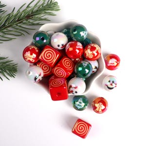 May include: A white bowl filled with red, green and white Christmas themed beads. The beads are round and square with various designs including snowmen, gingerbread men and swirls.