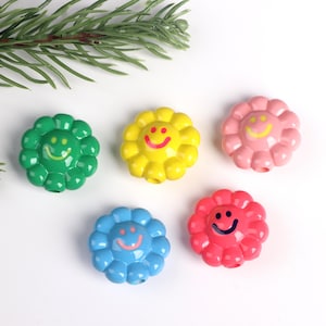 Peut inclure: Cinq perles en forme de fleurs souriantes, de couleurs verte, jaune, rose, bleue et rouge. Chaque perle est percée d'un trou pour l'enfilage. Un brin de feuillage vert est en arrière-plan.