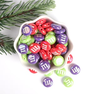 Pu&ograve; includere: Una ciotola piena di perline rotonde colorate con la lettera "M" stampata su di esse. Le perline sono in tonalit&agrave; di verde, rosso e viola. Alcune perline sono sparse fuori dalla ciotola.