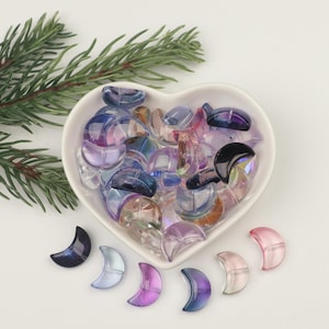 May include: A heart-shaped bowl filled with iridescent crescent moon-shaped beads in shades of purple, blue, and pink. The beads are scattered around the bowl and on a white surface.