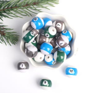 Può includere: Una ciotola piena di perline di plastica a forma di palla da biliardo colorate. Le perline sono bianche, blu, verdi e grigie con numeri neri su di esse. Le perline sono in una ciotola bianca con un bordo a forma di conchiglia.