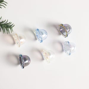 May include: Seven iridescent glass bell charms in various colours, including blue, white, and purple. The charms have a scalloped edge and a rounded top.