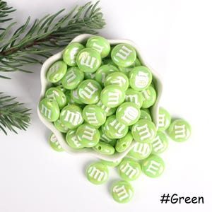 Pu&ograve; includere: Una ciotola piena di perline di plastica verde e bianca. Ogni perlina ha la lettera "M" stampata su di essa. Le perline sono disposte in uno schema casuale. #Verde