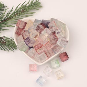 May include: A bowl filled with colorful, square-shaped glass beads. The beads have a frosted, translucent appearance and are decorated with intricate patterns. Some of the beads are scattered around the bowl.