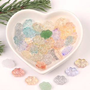 Puede incluir: Un bol lleno de cuentas de vidrio translúcidas en forma de flor de colores. Las cuentas son de vidrio y tienen una variedad de colores, incluyendo rosa, azul, verde y amarillo. Las cuentas están dispersas alrededor del bol y algunas están sobre la superficie blanca.