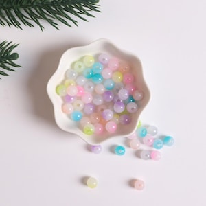 May include: A white bowl filled with pastel-colored round beads. The beads are translucent and have a slight iridescent shimmer. Some beads have spilled out of the bowl and are scattered on the white surface.
