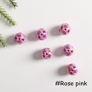 May include: Six pink round beads with a black and white pattern. The beads have a hole in the center and are scattered on a white surface. #Rose pink