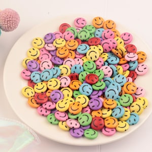 May include: A collection of colorful smiley face beads in various colors, including yellow, orange, green, blue, pink, purple, and red. The beads are arranged on a white plate, creating a cheerful and playful aesthetic. These beads are ideal for crafting and jewelry making.