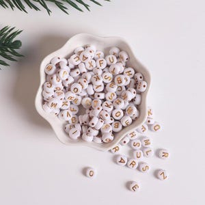 Peut inclure: Un bol blanc rempli de perles blanches en alphabet avec des lettres dorées. Les perles sont dispersées autour du bol.