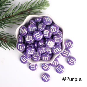 Pu&ograve; includere: Perle di colore viola con la lettera "M" in bianco in una ciotola bianca. Le perle sono iridescenti e hanno una lettera "M" bianca su ciascuna di esse. #Purple