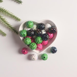 May include: A white heart-shaped bowl filled with colorful beads. The beads are green, pink, black, and white. They have a swirl design.