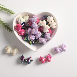 May include: A heart-shaped bowl filled with iridescent plastic bow-shaped beads in shades of pink, purple, and white. The beads are scattered on a white surface.