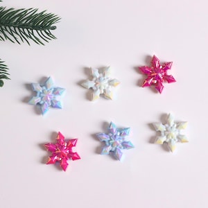 Könnte beinhalten: Sechs schimmernde Schneeflocken-Ornamente in verschiedenen Blautönen und Rosa. Die Schneeflocken sind auf einem weißen Hintergrund angeordnet.
