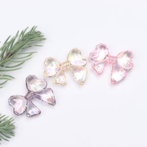 May include: Three iridescent plastic bows in different shades of pink, purple, and yellow. The bows are arranged in a row on a white surface.