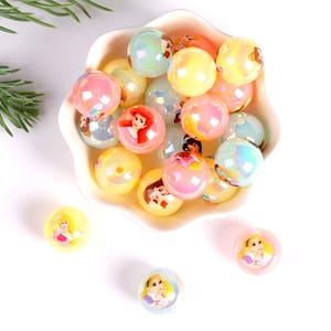 May include: A white bowl filled with iridescent pastel-colored beads featuring princess characters. The beads are approximately 2 cm in diameter.