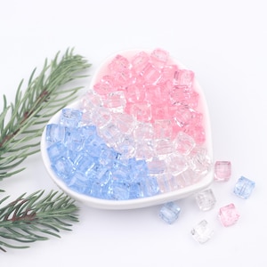 Könnte beinhalten: Eine herzförmige Schale gefüllt mit kleinen, quadratischen, durchsichtigen Perlen in den Farben Rosa, Blau und Klar. Die Perlen sind um die Schale herum verstreut.