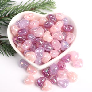 May include: A white heart-shaped bowl filled with iridescent pink and purple heart-shaped beads. The beads are scattered around the bowl on a white surface.