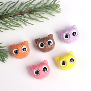 May include: Five colorful cat-shaped buttons with googly eyes. The buttons are orange, brown, purple, yellow, and pink.