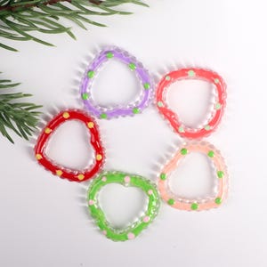 May include: A collection of colorful, clear plastic teething rings in various shapes, including a triangle and a heart. Each ring is decorated with small, colorful dots. The rings are arranged on a white surface, with a sprig of greenery in the background.