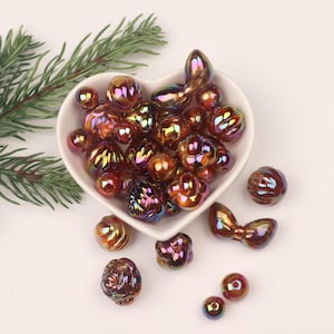 Puede incluir: Un bol con forma de corazón lleno de cuentas de vidrio marrón y morado iridiscentes. Las cuentas son de varias formas, incluyendo redonda, ovalada y de lágrima. Algunas cuentas tienen una superficie texturizada. También hay varias cuentas esparcidas alrededor del bol.