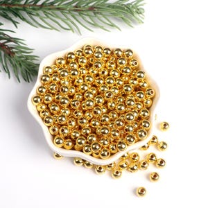 May include: A white bowl filled with shiny gold round beads. The beads are scattered around the bowl on a white surface.