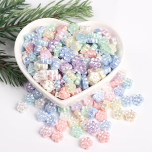 May include: A heart-shaped bowl filled with pastel-colored flower-shaped beads. The beads are scattered around the bowl on a white surface.