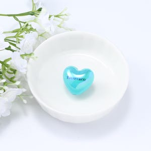 Könnte beinhalten: Ein herzförmiges, türkisfarbenes Schmuckstück mit dem Text "TIFFANY&CO" in Weiß. Es liegt in einer flachen, weißen Schale. Weiße Blumen und Grünzeug befinden sich im Hintergrund. Das Schmuckstück ist wahrscheinlich aus Glas.