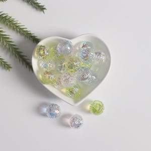 May include: A white heart-shaped dish filled with iridescent glass beads in shades of green and blue. The beads have a textured surface and are scattered randomly in the dish.