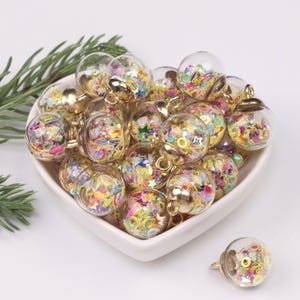 Puede incluir: Una colección de colgantes de globo de cristal transparente llenos de confeti colorido. Cada colgante tiene una tapa y un lazo de metal dorado para colgar. Los colgantes se muestran en un plato blanco en forma de corazón.