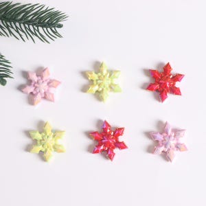 Könnte beinhalten: Sechs schimmernde Schneeflocken-Ornamente in verschiedenen Farben, darunter rosa, gelb und rot. Die Schneeflocken sind symmetrisch auf einem weißen Hintergrund angeordnet.