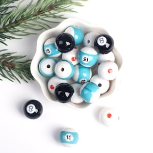 May include: A white bowl filled with ceramic billiard ball beads. The beads are blue, white, and black with numbers 8 and 15 printed on them. Some beads have a red dot on them.