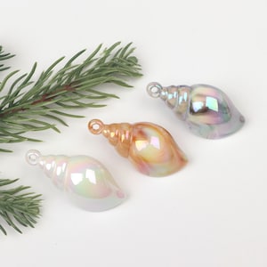 Può includere: Tre ciondoli a forma di conchiglia iridescente con un foro nella parte superiore per appendere. Le conchiglie sono in tonalità di bianco, arancione e viola.