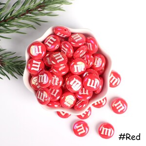 Pu&ograve; includere: Una ciotola piena di perline rosse a forma di caramella con una "M" bianca su ogni perlina. Le perline sono sparse intorno alla ciotola su una superficie bianca. #Rosso