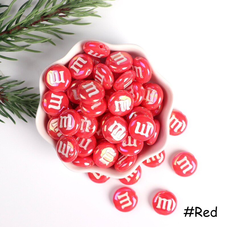 Pu&ograve; includere: Una ciotola piena di perline rosse a forma di caramella con una "M" bianca su ogni perlina. Le perline sono sparse intorno alla ciotola su una superficie bianca. #Rosso