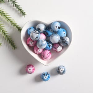 May include: A heart-shaped bowl filled with colorful, marbled beads. The beads are a mix of blue, pink, and white with black swirls. There are also a few beads outside of the bowl.